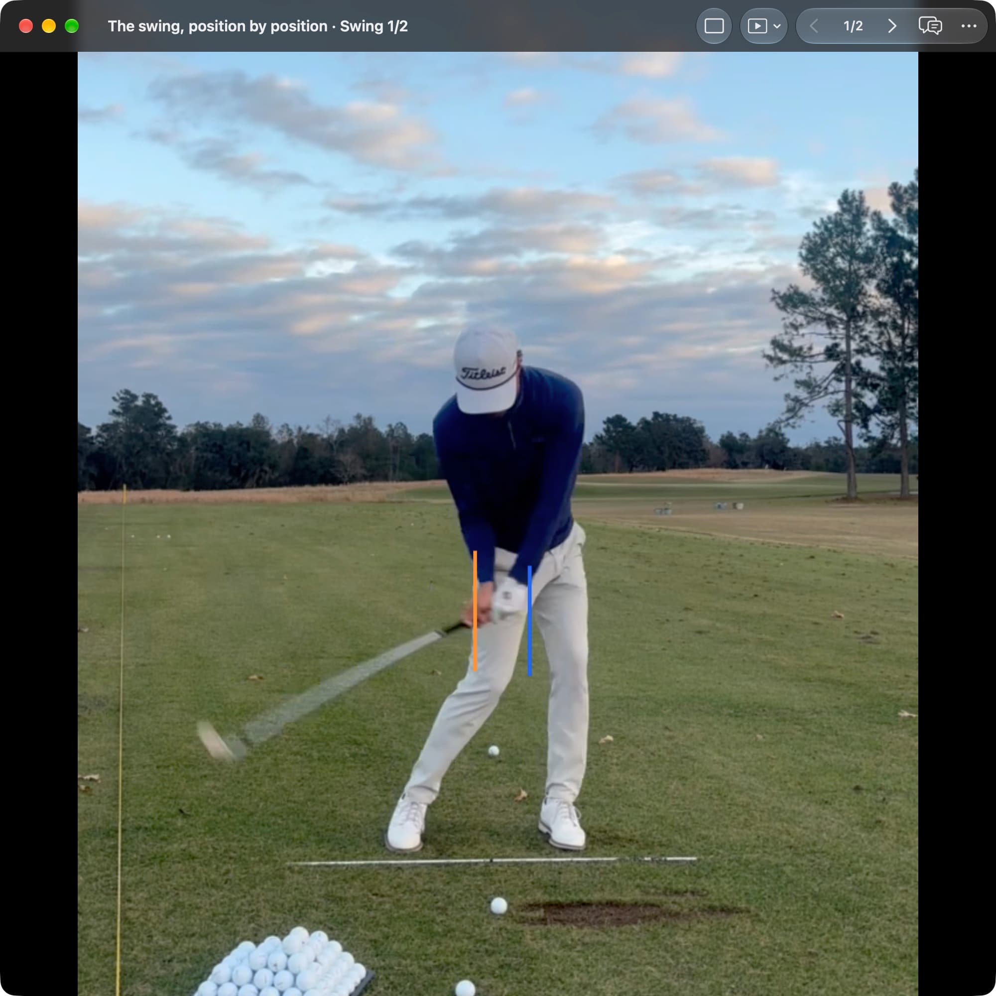 A golfer in MotionEdge Video Player, face-on on a range, just before impact with the club blurred behind the hands. An orange line marks the pelvis center at Address and a blue line sits clearly toward the target.