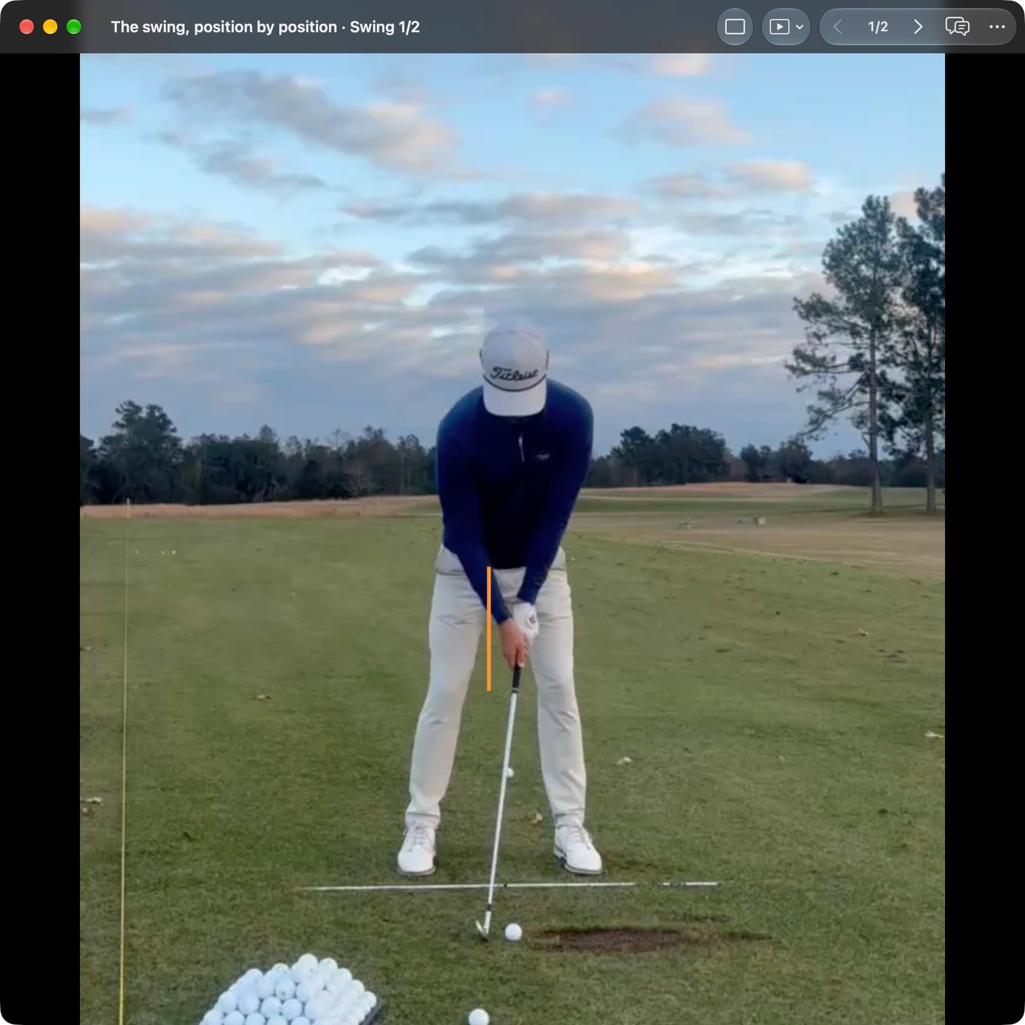 A golfer at Address in MotionEdge Video Player, face-on on a range. An orange vertical line marks the center of the pelvis. The ball sits between the feet near an alignment stick, and the handle is slightly ahead of the clubhead.